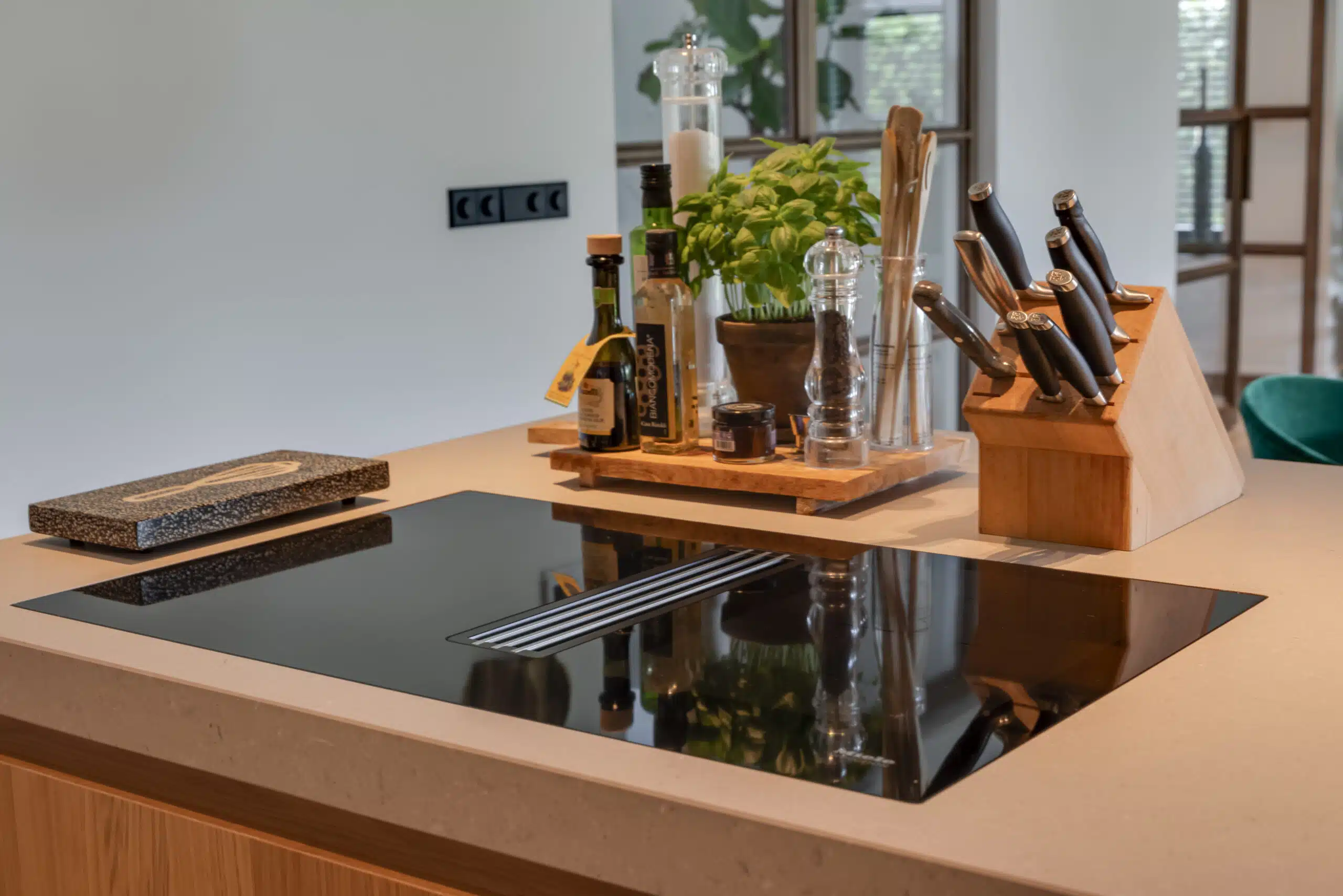 Detail van een luxe keuken in Hendrik-Ido-Ambacht met een inductiekookplaat geïntegreerd in een composiet werkblad in beige tinten. Op het kookeiland staan stijlvolle accessoires zoals kruidenpotjes, olijfolieflessen, een houten messenblok en een verse basilicumplant